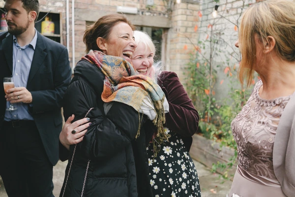 Two women laughing freely among wedding guests in a brick garden, much joy in the air