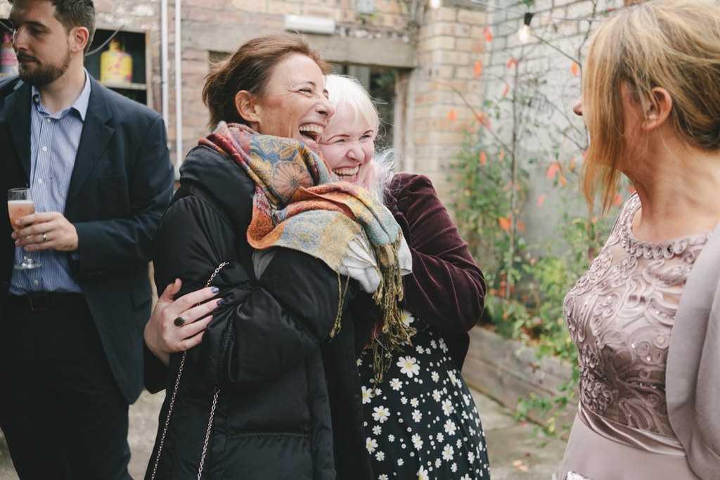 Two women laughing freely among wedding guests in a brick garden, much joy in the air
