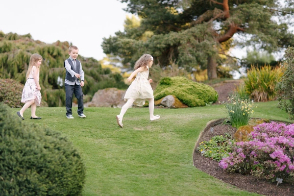 Three children in formal wedding attire run and play on the lawn of the Royal Botanic Garden Edinburgh. Candid documentary wedding moment full of movement and energy.
