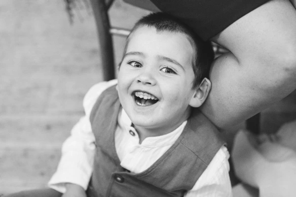 A young boy in a waistcoat laughs with his mouth wide open at the wedding reception, leaning back against an adult. Black and white candid documentary portrait.