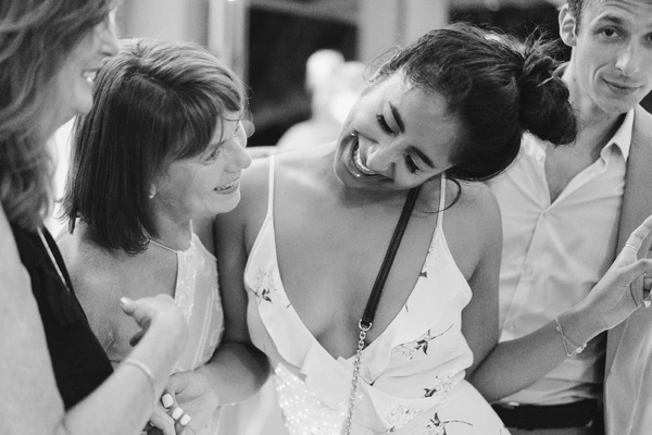 Bride laughing happily among guests in a floral white dress, natural light at the reception