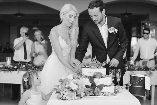 Newlyweds cut the cake as a little girl watches intently — guests, floral decoration in background