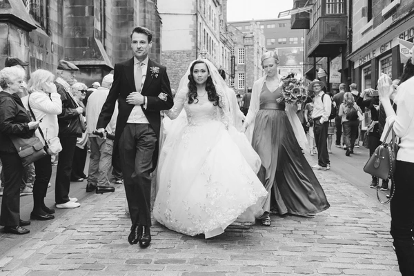Bride and groom walk along a city street surrounded by guests — white lace, veil, classic black-and-white
