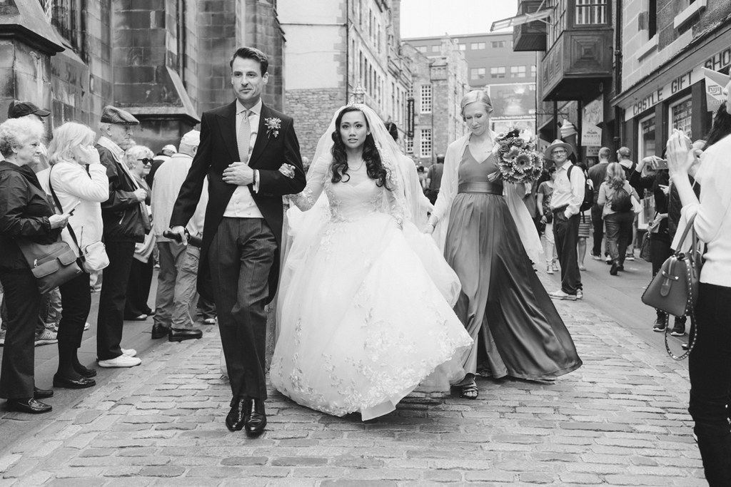 Bride and groom walk along a city street surrounded by guests — white lace, veil, classic black-and-white