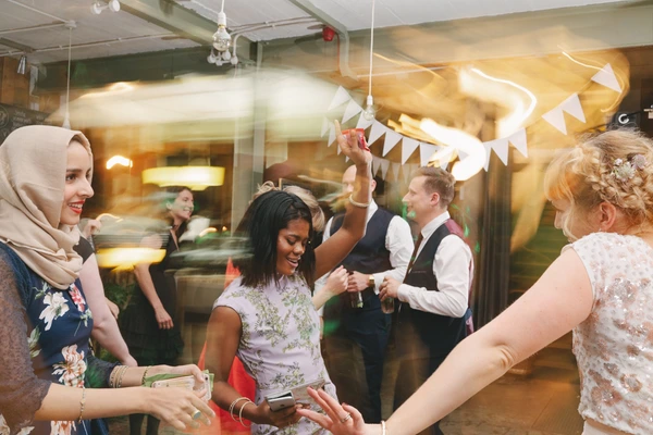 Guests dance and laugh at the wedding reception under white bunting and warm lamp lighting
