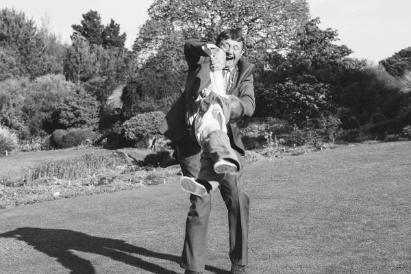 Black and white image of a laughing man in a suit holding a young child upside down on a garden lawn. Candid and joyful documentary wedding moment.
