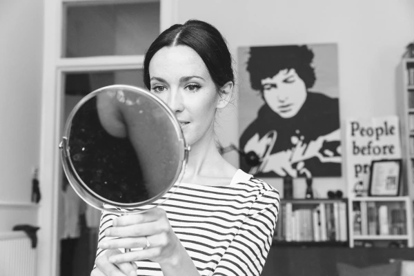 Black and white portrait of the bride holding a round mirror up toward the camera with a Bob Dylan poster visible in the background. Playful moment during preparations.