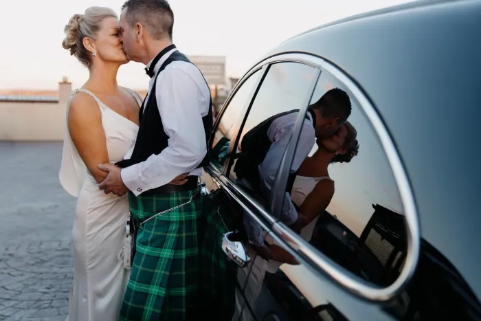 Bride and groom kissing beside black car at sunset, groom wearing green tartan kilt, couple's reflection visible in car window.