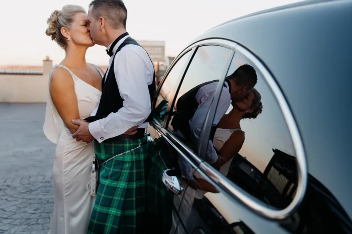 Bride and groom kissing beside black car at sunset, groom wearing green tartan kilt, couple's reflection visible in car window.