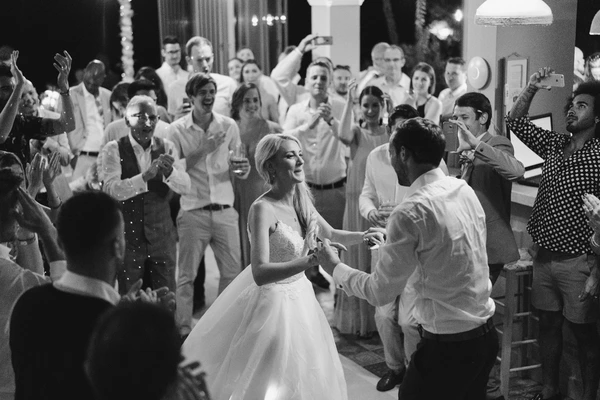 Bride in white dances with groom on the dance floor, joyful guests clapping and celebrating at night