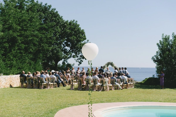 Ceremony on a green lawn overlooking the sea — white balloon above pool, guests seated