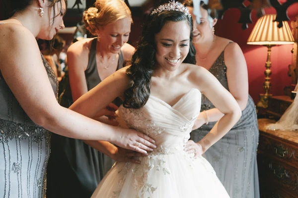 Bride in white lace with tiara, smiling as bridesmaids surround and smooth her gown