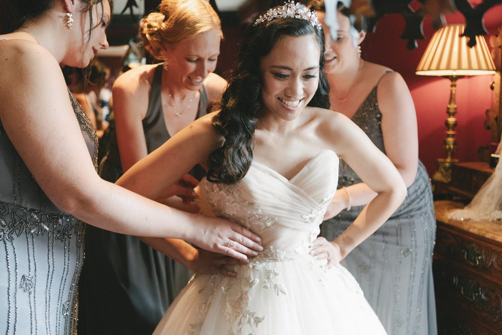 Bride in white lace with tiara, smiling as bridesmaids surround and smooth her gown