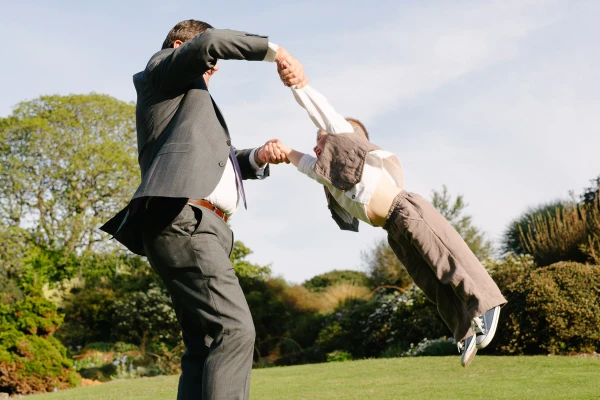 A man in a suit whirls a small boy through the air by his arms on a sunny garden lawn. Candid documentary wedding photography capturing playful family moments.