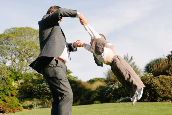 A man in a suit whirls a small boy through the air by his arms on a sunny garden lawn. Candid documentary wedding photography capturing playful family moments.