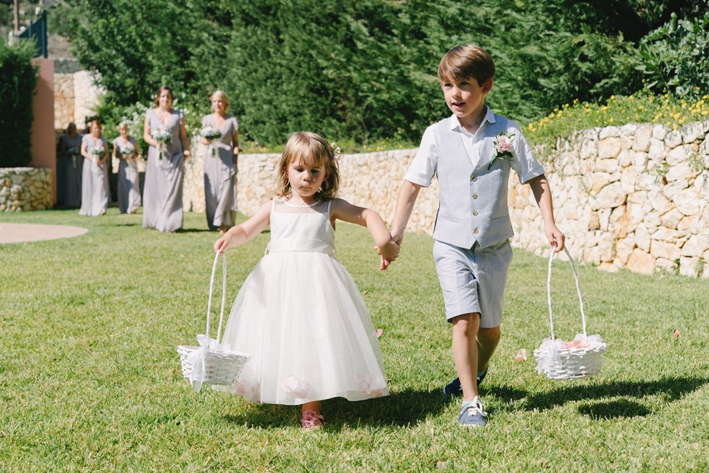Two children with flower basket walk in the garden at the wedding — bride and father in background