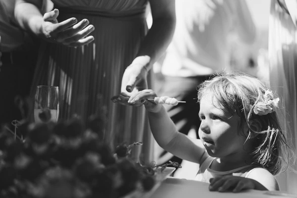 Little girl with floral hair clip puts finger to her mouth — guests smile at the curious moment. B&W.
