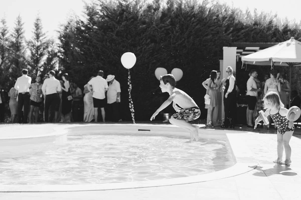 Woman and child play at the poolside in the evening, guests watching — fairy lights in garden