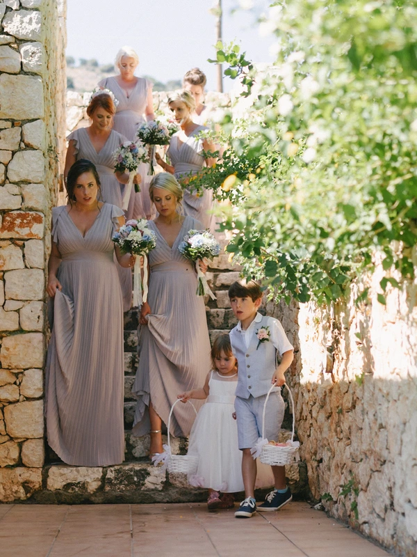 Bridesmaids and flower girls walk through a narrow ivy-covered stone lane, bouquets in hand