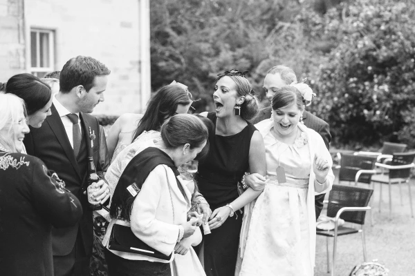 Black and white image of a group of wedding guests laughing together outside, one woman with an open-mouthed expression of surprise. Candid documentary wedding photograph.