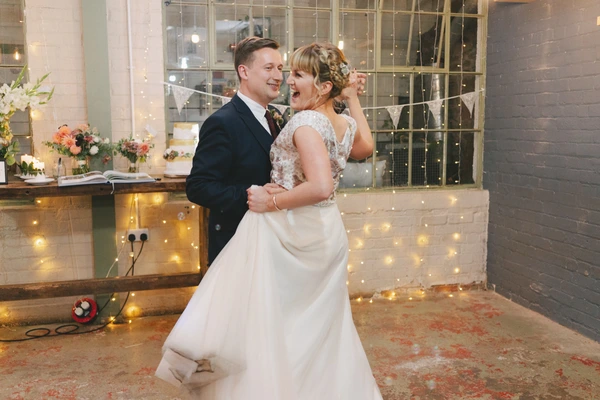 Joyful bride and groom dancing in a glass reception room decorated with warm fairy lights