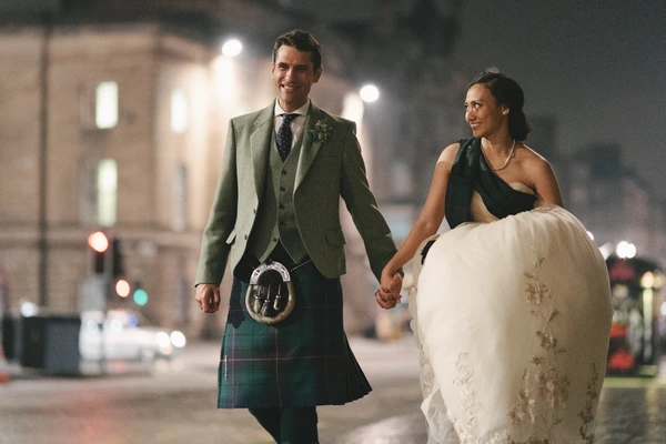 Groom in Scottish kilt and green blazer, bride in white lace — walking hand in hand at night, smiling