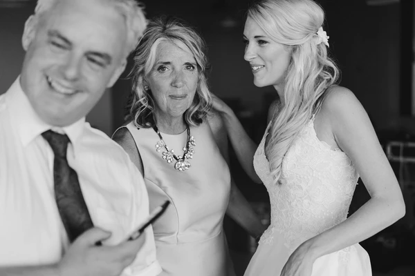 Bride in white lace smiles talking to an older woman and a man in white shirt — black-and-white