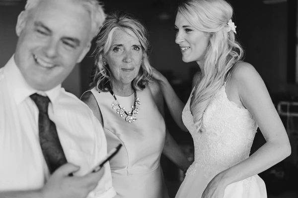 Bride in white lace smiles talking to an older woman and a man in white shirt — black-and-white