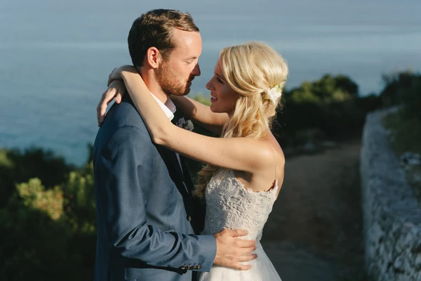 Young couple embrace at a castle wall — white lace dress, dark blue suit, sea and green hills behind
