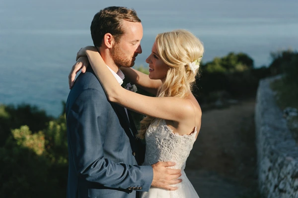 Young couple embrace at a castle wall — white lace dress, dark blue suit, sea and green hills behind