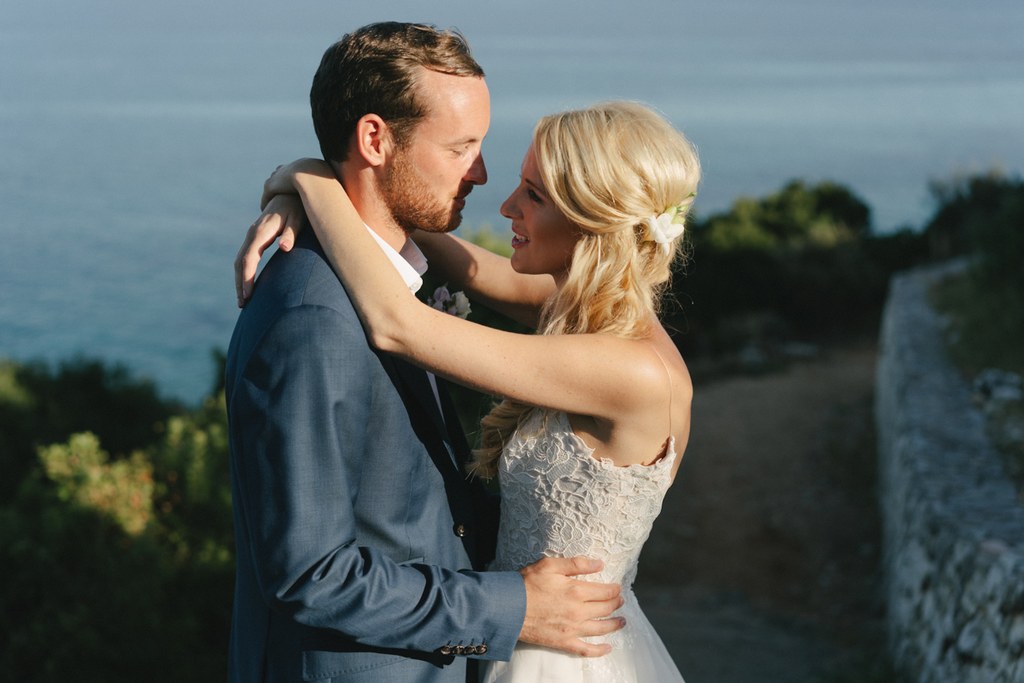 Young couple embrace at a castle wall — white lace dress, dark blue suit, sea and green hills behind