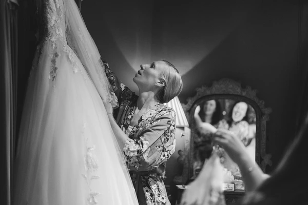 Bride admires her lace gown while getting dressed, friends preparing for the wedding in background