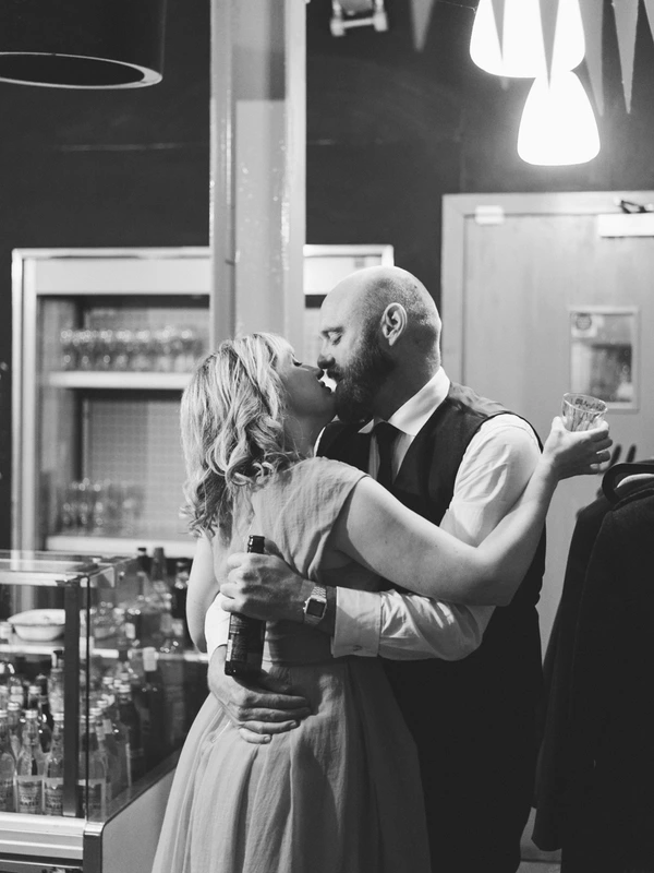 Wedding kiss — groom in white shirt approaches the smiling bride, warm lamp lighting at the bar