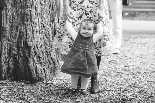 Black and white candid shot of a toddler in a pinafore dress standing and holding an adult's hand against a large tree trunk after the ceremony. Sweet documentary moment.