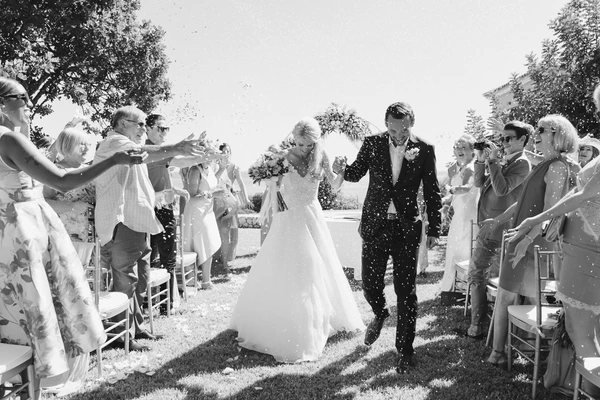 Bride and groom leave the altar, confetti falling around them — black-and-white, green lawn