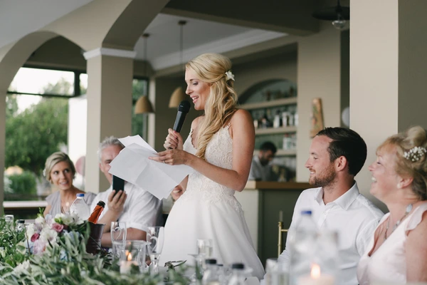 Bride with microphone addresses guests at the dinner, smiling, reading from paper — family watching