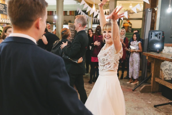 Bride in white lace top dances joyfully with the groom, guests watching with smiles