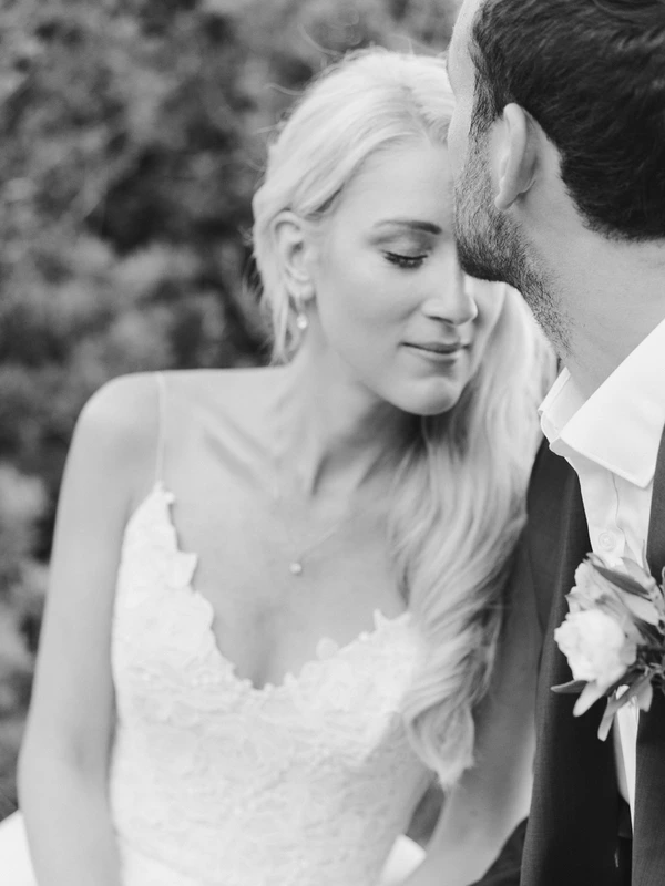 Bride with long wavy blonde hair smiles, groom embraces her — green bushes behind. Black-and-white.