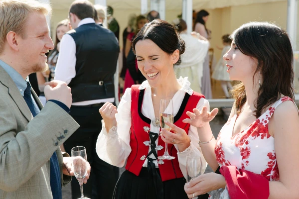 Candid shot of the bride in her Norwegian bunad laughing and gesturing animatedly while chatting with guests at the outdoor drinks reception, champagne in hand. Joyful documentary moment.