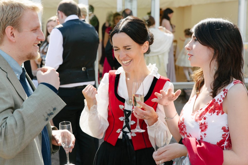 Candid shot of the bride in her Norwegian bunad laughing and gesturing animatedly while chatting with guests at the outdoor drinks reception, champagne in hand. Joyful documentary moment.