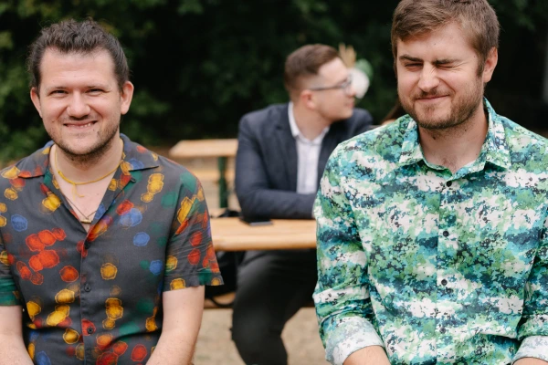 Two men in colorful patterned shirts sitting outdoors, one smiling at the camera, one squinting.