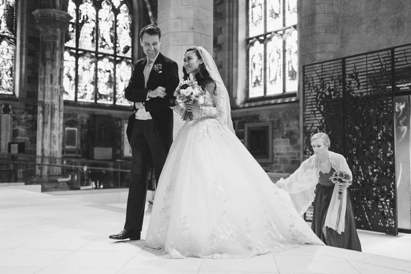 Bride and groom walk past church choir, bridesmaid carrying the lace veil — natural light
