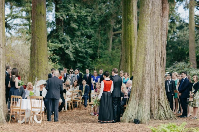 Outdoor woodland wedding ceremony with guests seated among tall trees, bride in red bodice and groom facing officiant