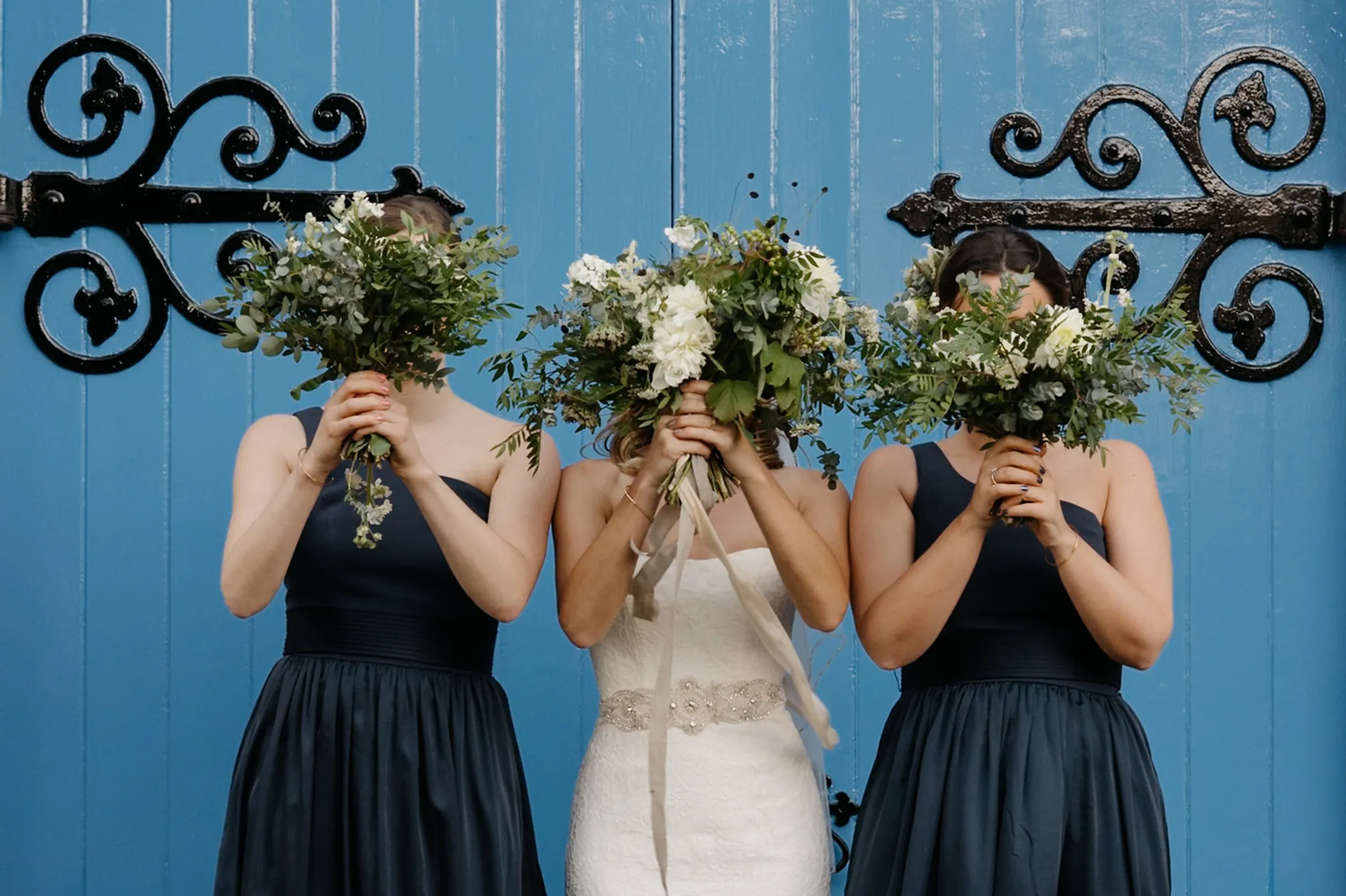 Bride and two bridesmaids in navy dresses hide faces behind lush green and white bouquets against a blue door.