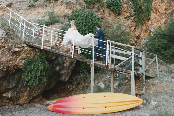Bride in white and groom in navy walk by the wooden bridge at the rocky shore, surfboard on the sand