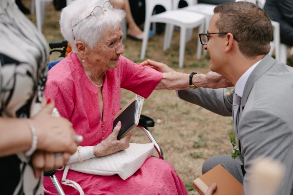 Groom talking to elderly guest in pink dress during outdoor wedding reception on garden venue.