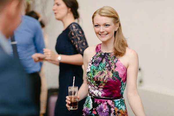 A smiling woman in a colourful floral dress holds a drink with a paper straw at the wedding reception, looking warmly toward the camera. Candid documentary wedding portrait.