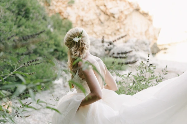 Bride from behind in white tulle gown with floral hair wreath on rocky terrain among green plants