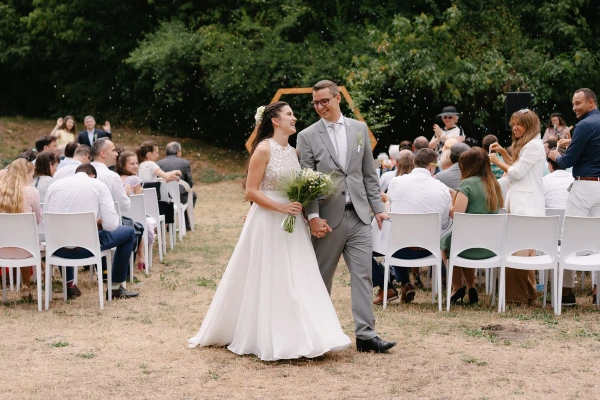 Bride and groom smiling walking together after ceremony with guests seated in background garden setting.