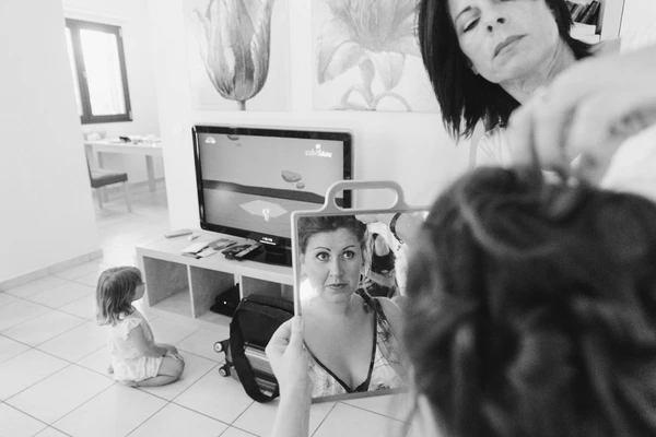 Bride sits at a mirror as makeup artist works — little girl watching in background. Black-and-white.